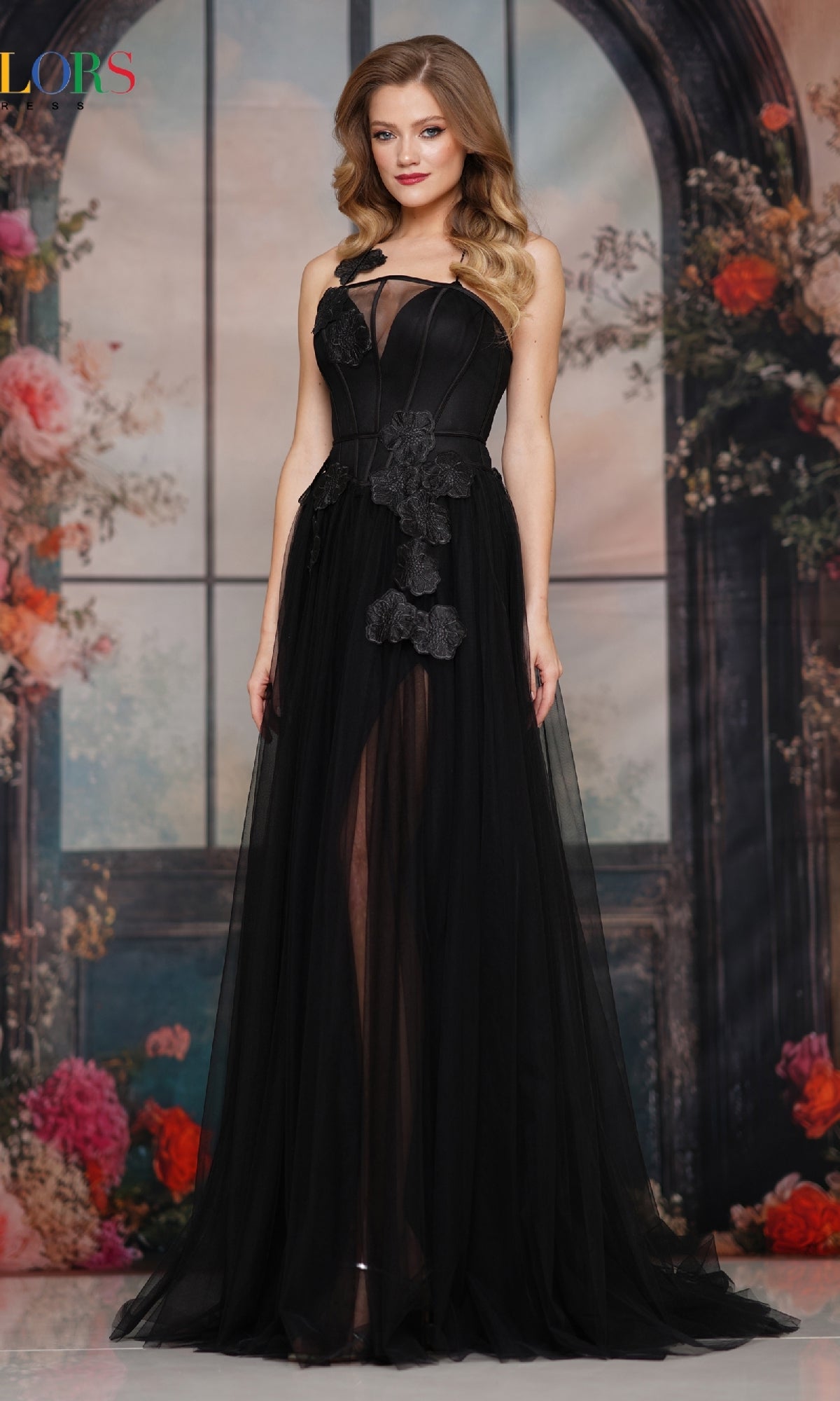 A woman wearing the Colors Dress 3796, a long prom dress with an a-line tulle skirt and 3D floral appliques, stands in front of a floral and windowed backdrop.